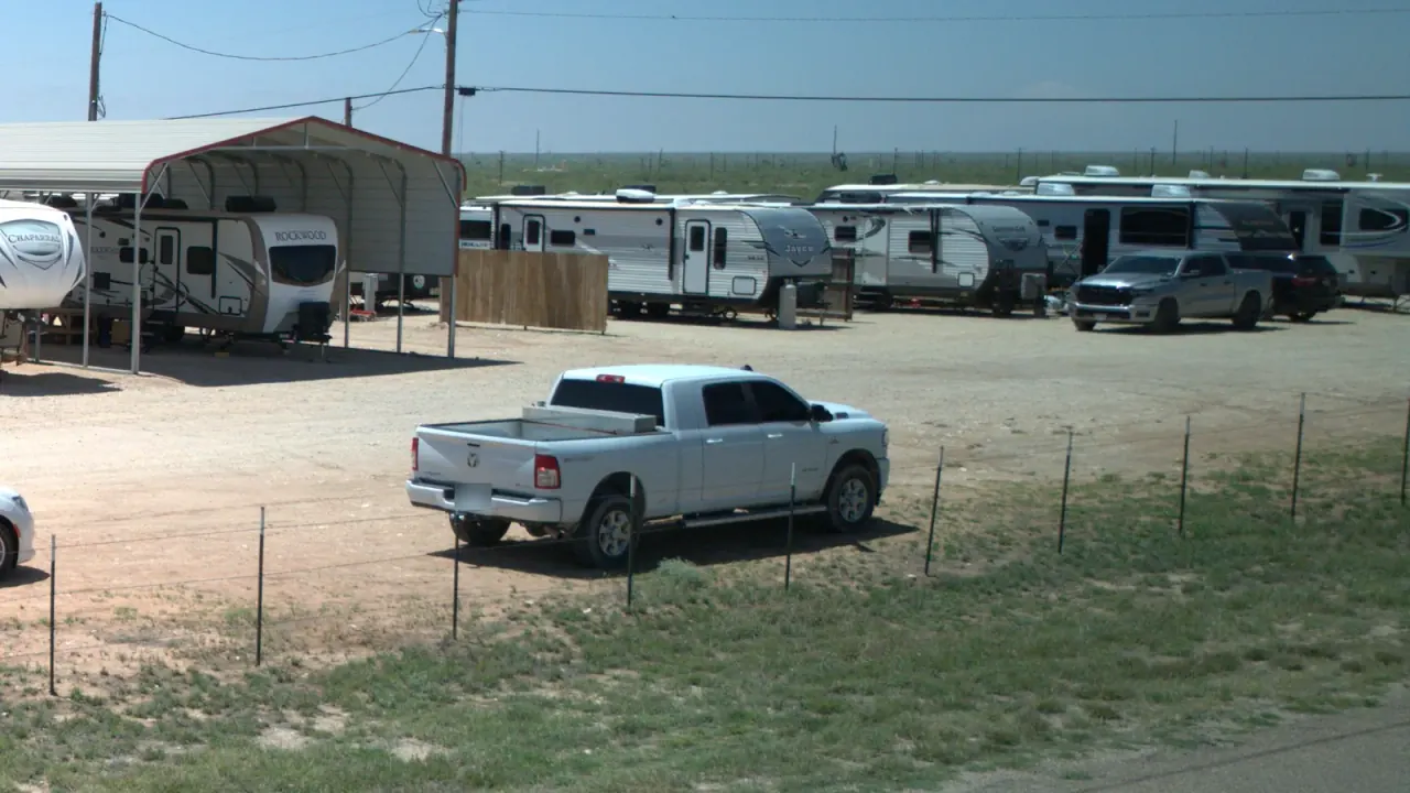 RVs and a white pickup truck parked on gravel at Goldsmith RV Park & Housing, seen from the road, with a metal carport canopy and a wire fence along the front