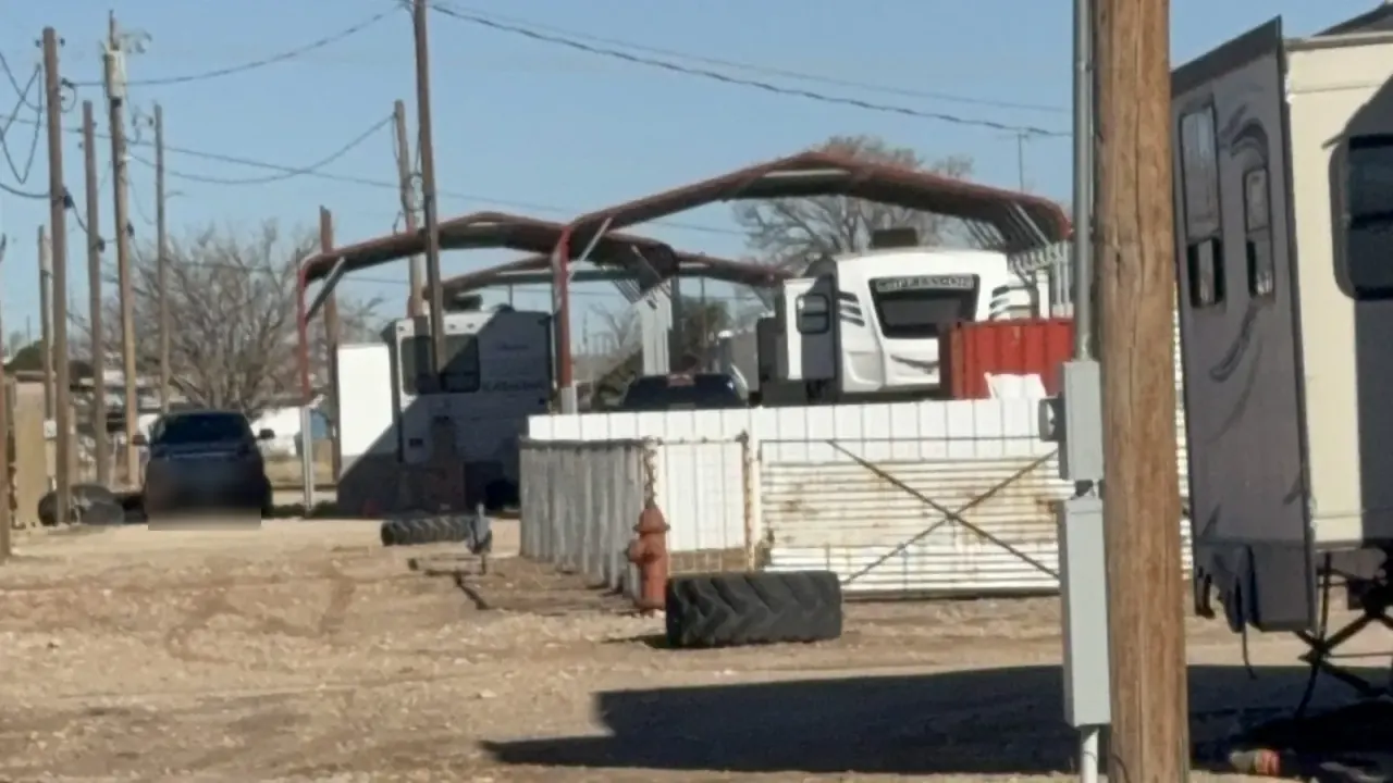 RVs, a pickup truck and metal carport canopies on gravel at Goldsmith RV Park & Housing on a clear winter day, with bare trees and power poles