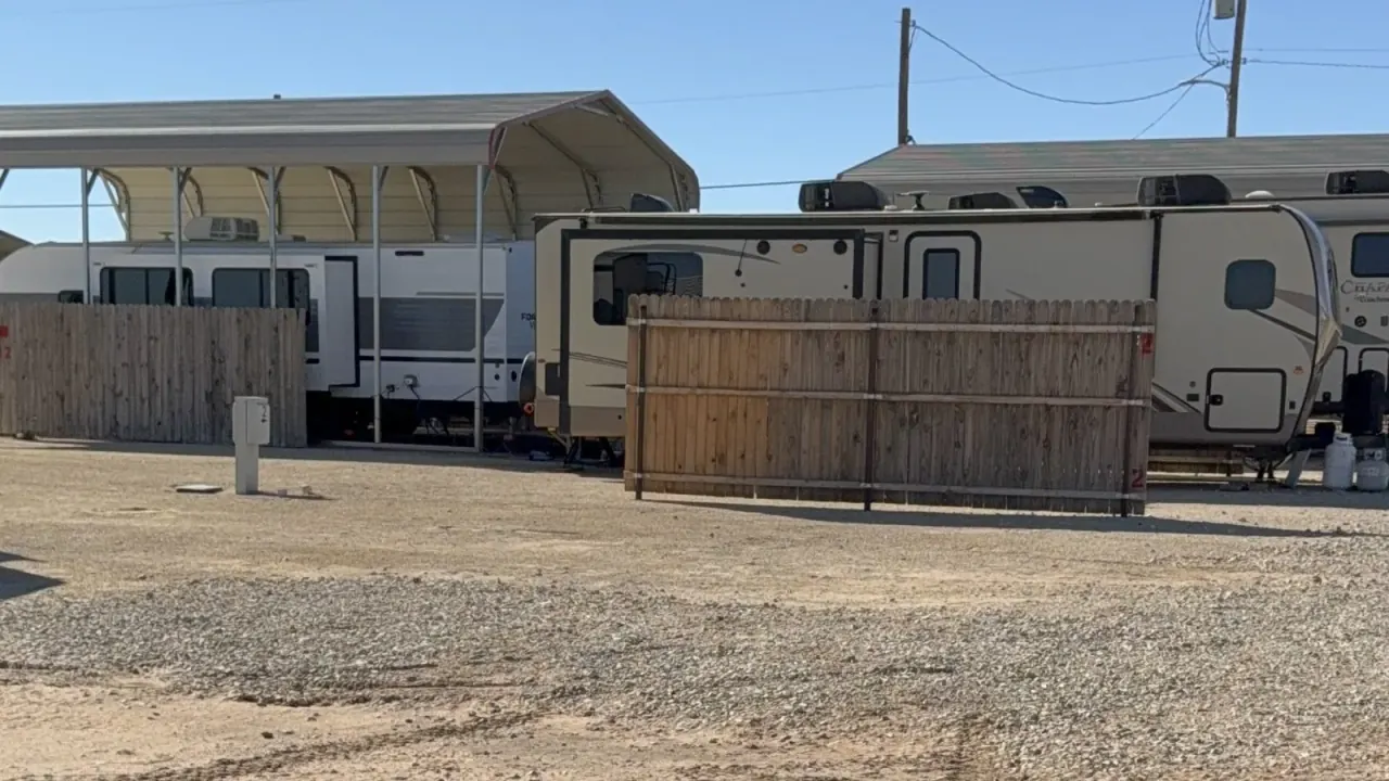 Travel trailers parked on gravel at Goldsmith RV Park & Housing, with wooden privacy fences between the sites, a metal carport canopy over one trailer, and a small white utility post in the foreground
